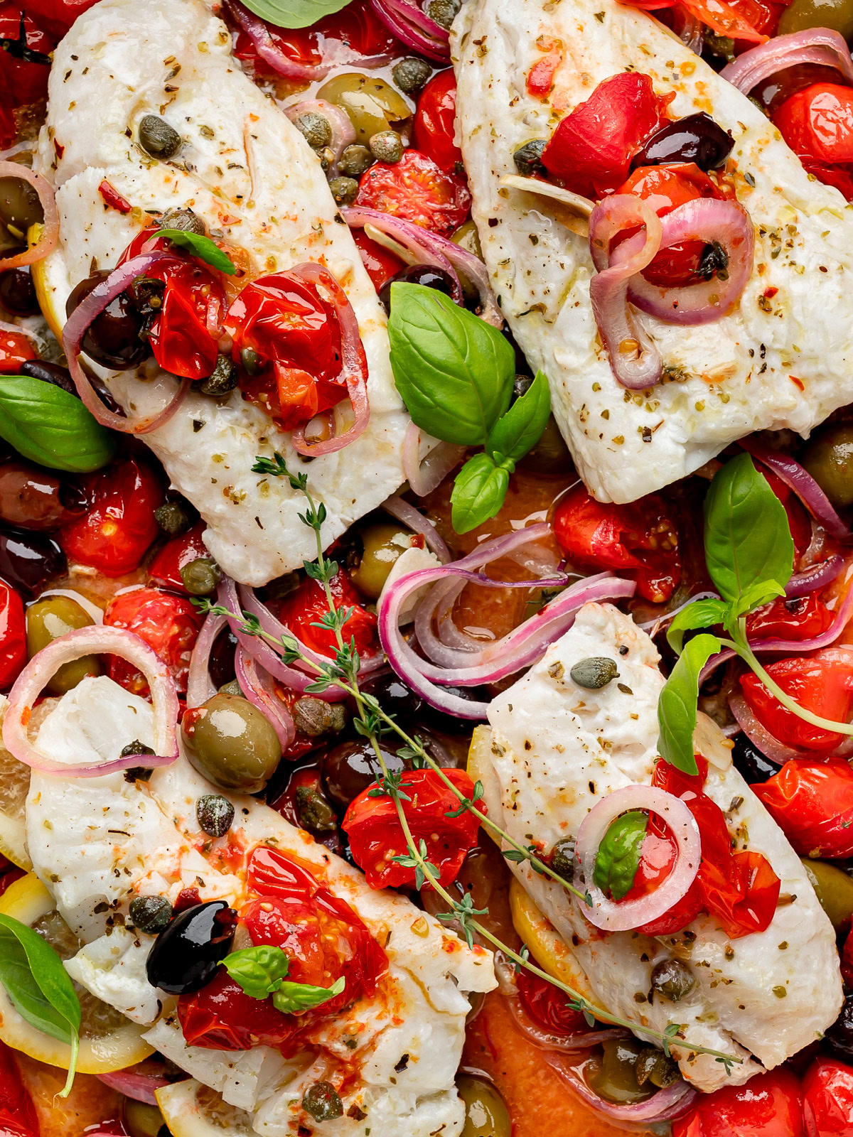 Close-up of baked cod fillets with cherry tomato mixture in baking dish, garnished with basil and thyme sprigs.