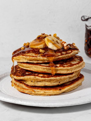 A stack of 4 pancakes on a white plate topped with sliced bananas, walnut and drizzles of maple syrup.