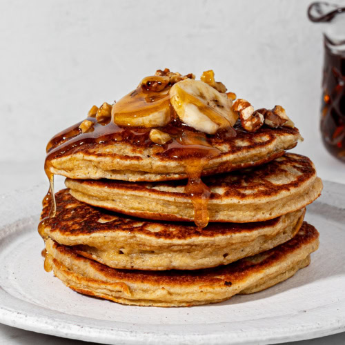 A stack of 4 pancakes on a white plate topped with sliced bananas, walnut and drizzles of maple syrup.