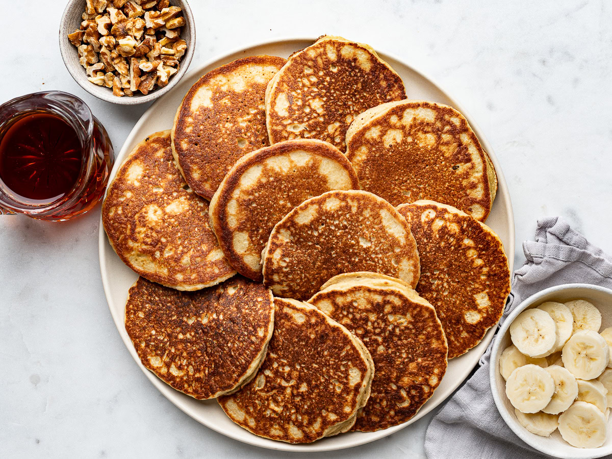 a large plate with cooked pancakes and a jar of maple syrup, walnuts and banana slices on the side.