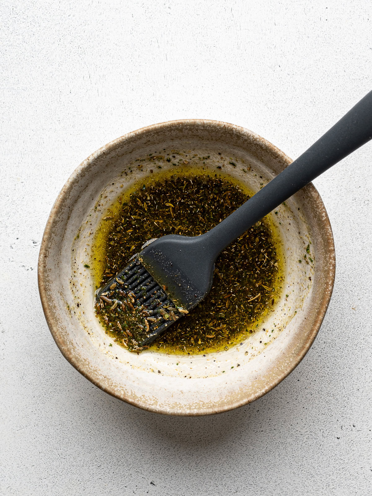 Olive oil and herb mixture in small bowl with silicone brush.