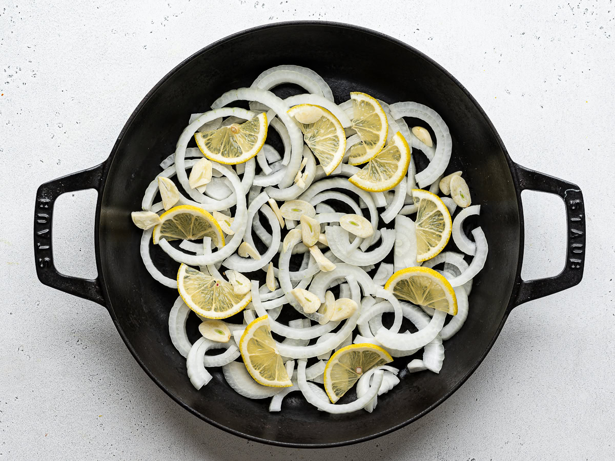 Sliced onions, lemon slices, and garlic slices in a black skillet.