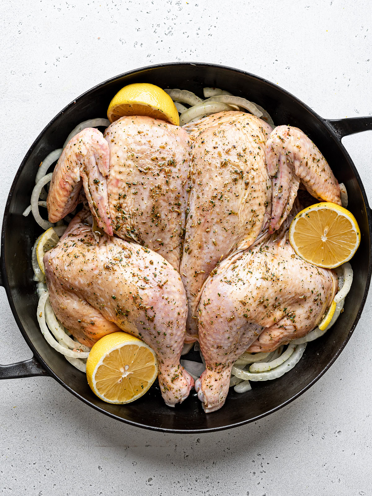 Un-cooked spatchcocked chicken in black skillet on top of onion mixture with lemon halves. 