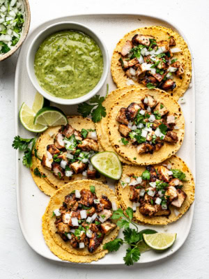 Five double layer corn tortillas on a white platter filled with chopped grilled chicken and topped with diced onion and chopped cilantro and a side of avocado salsa.