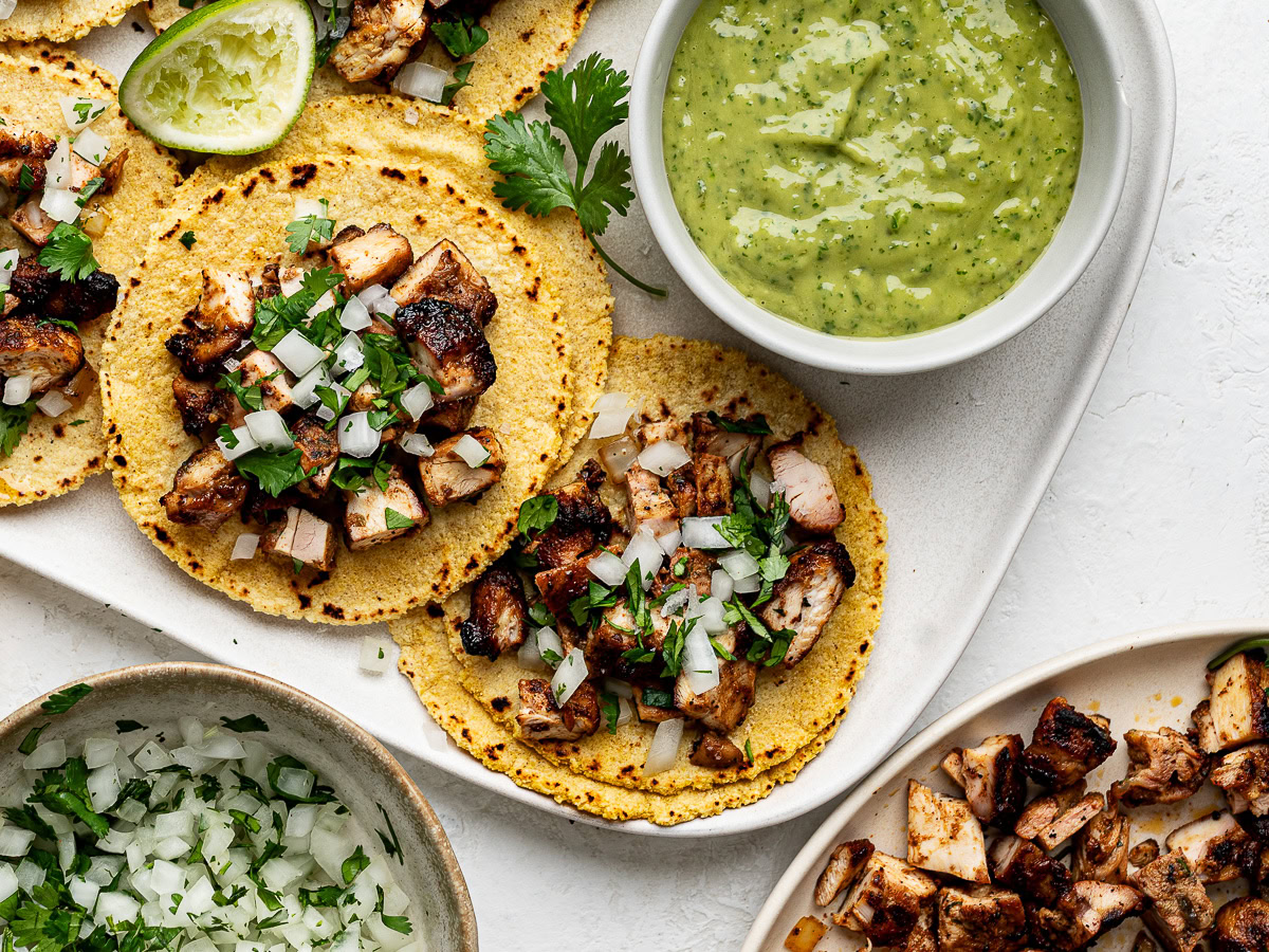Street chicken tacos on a white platter, avocado salsa, platter of chopped chicken, and a bowl of diced onion and chopped cilantro.