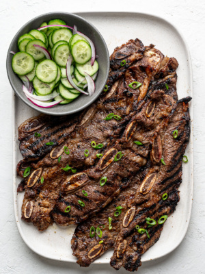 Grilled Korean-Style Short Ribs (Galbi) on a white rectangular platter with cucumber salad on the side.