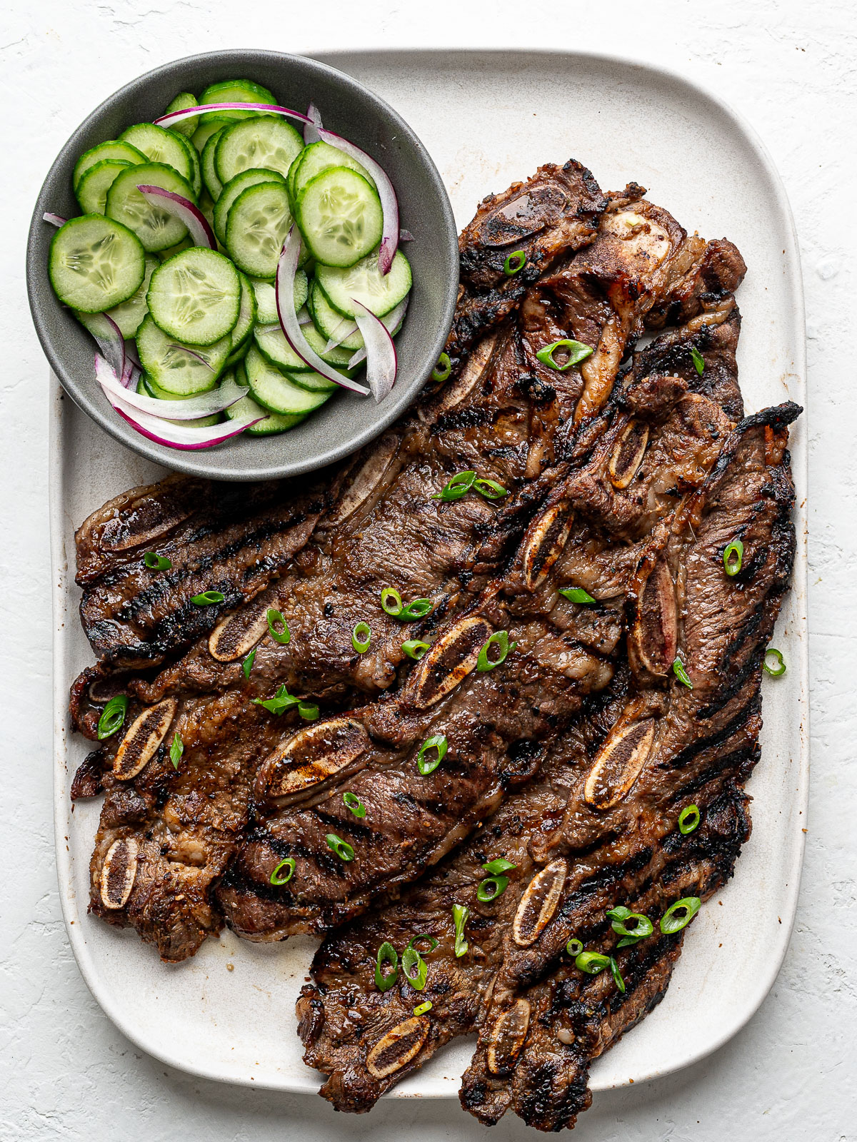 Grilled Korean-Style Short Ribs (Galbi) on a white rectangular platter with cucumber salad on the side.