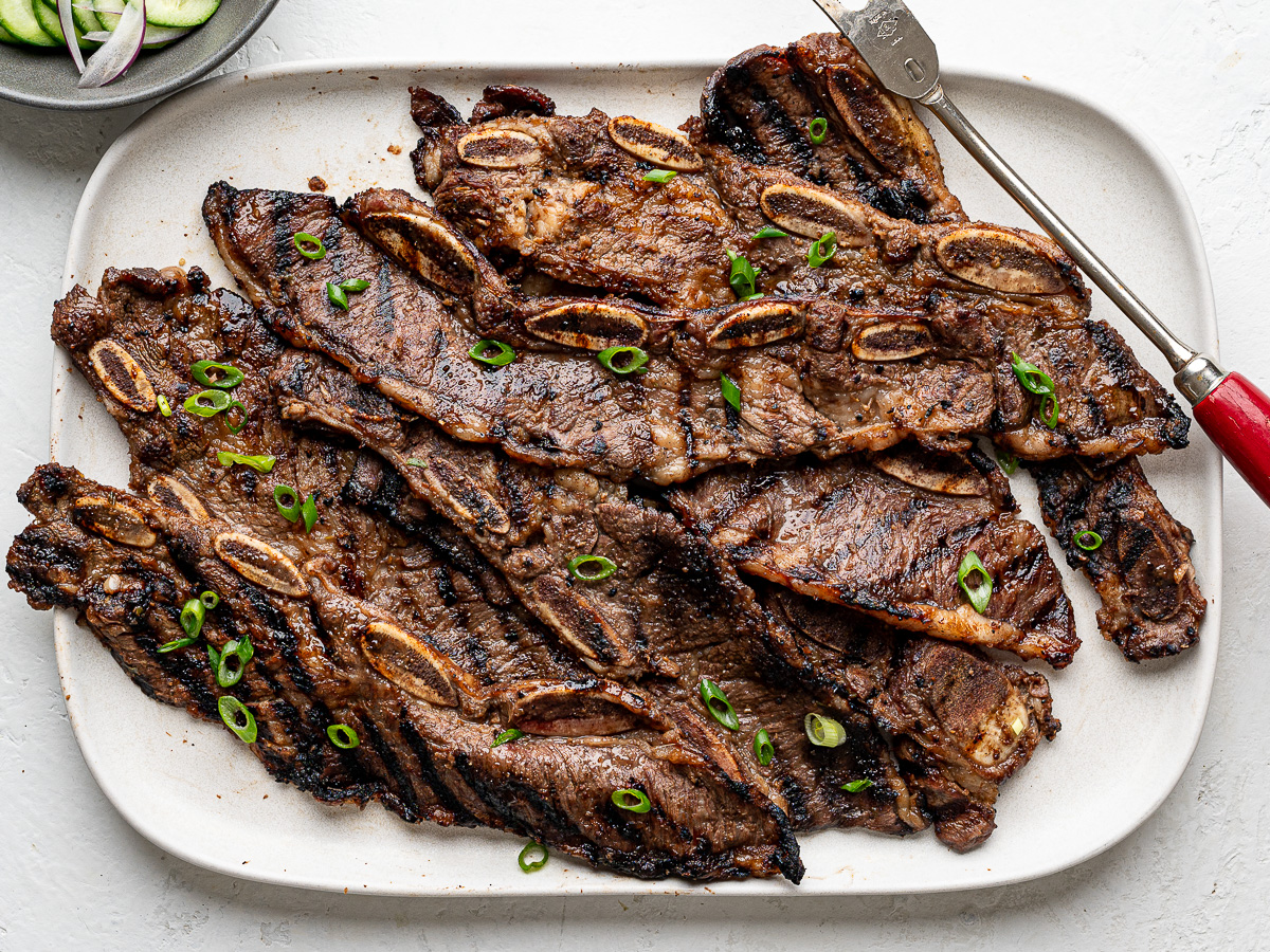 Grilled Short Ribs garnished with thinly sliced green onions on a white rectangular platter with cucumber salad on the side and large serving fork.