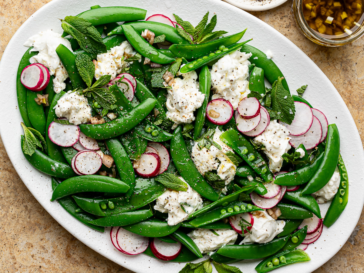 Close up of sugar snap pea and burrata salad served on an oval white platter with dressing on the side.