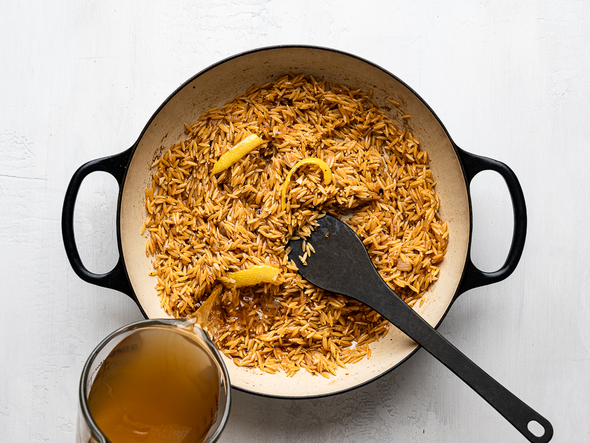 Pouring chicken broth into skillet with orzo mixture. 