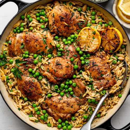Lemon Orzo topped with chicken thighs in black skillet and bowl of lemon slices on the side.