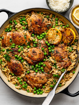 One-Pan Chicken And Lemon Orzo in black skillet with two charred lemon halves and a bowl of parmesan and a bowl of lemon slices on the side.