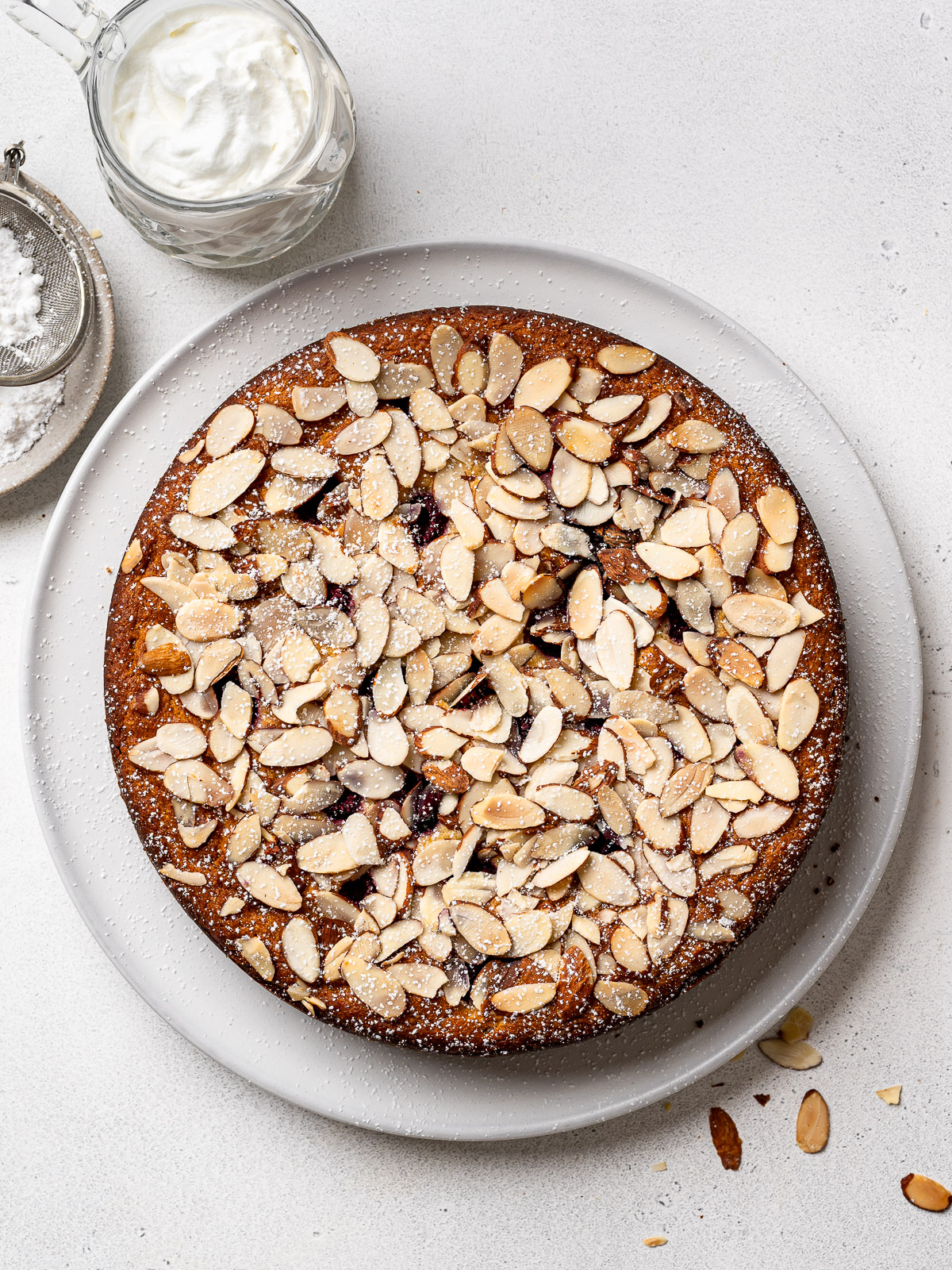 Raspberry Ricotta Almond Cake served on a white platter with a dusting of icing cuter and a jar of whipped cream on the side.