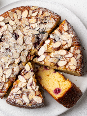Raspberry Almond Ricotta Cake on a white platter with one sliced piece flipped to show the inside crumb.
