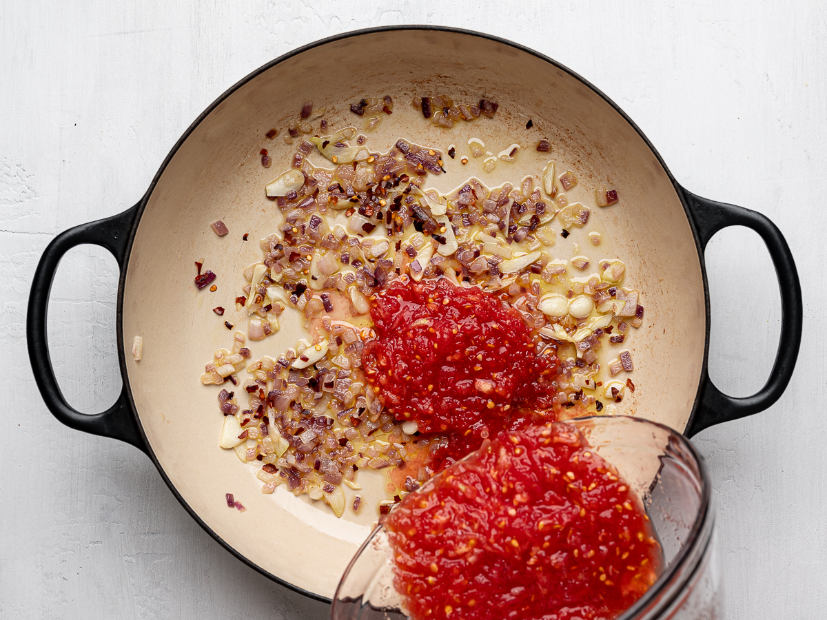 Pouring grated tomatoes into skillet with sautéed onion and garlic.