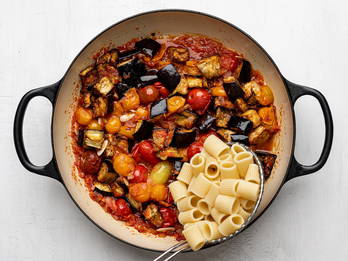 Adding cooked pasta with a skimmer to the skillet with eggplant mixture. 
