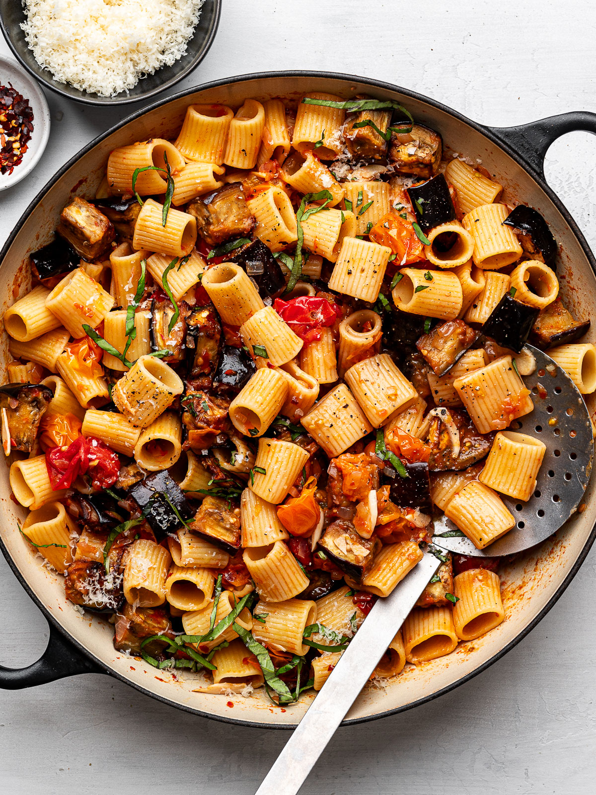 Roasted eggplant pasta in skillet with bowls of cheese and red pepper flakes on the side.