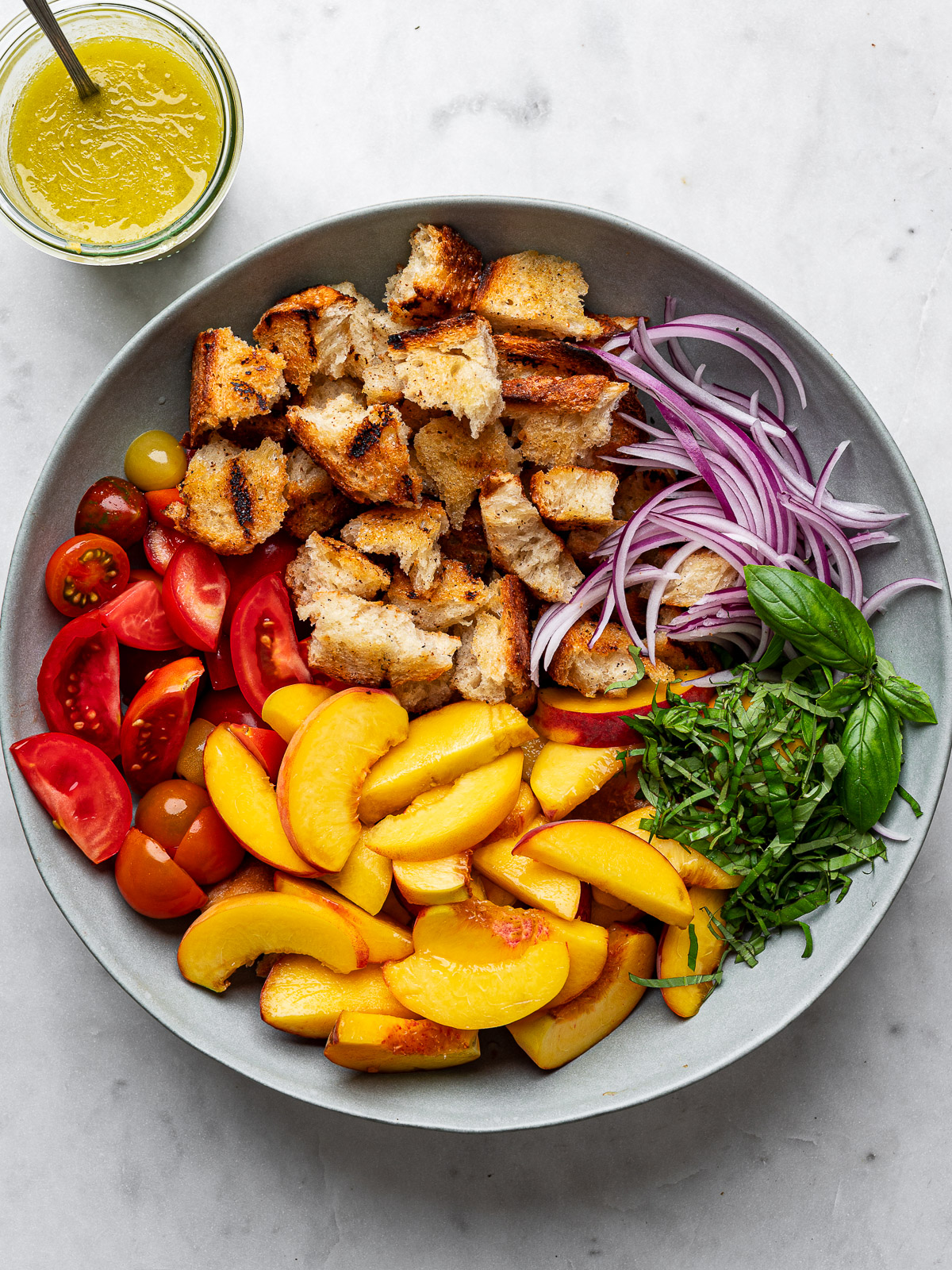 sliced peaches, tomatoes, onions, bread cubes, and basil arranged in a gray salad bowl with jar of vinaigrette on the side. 
