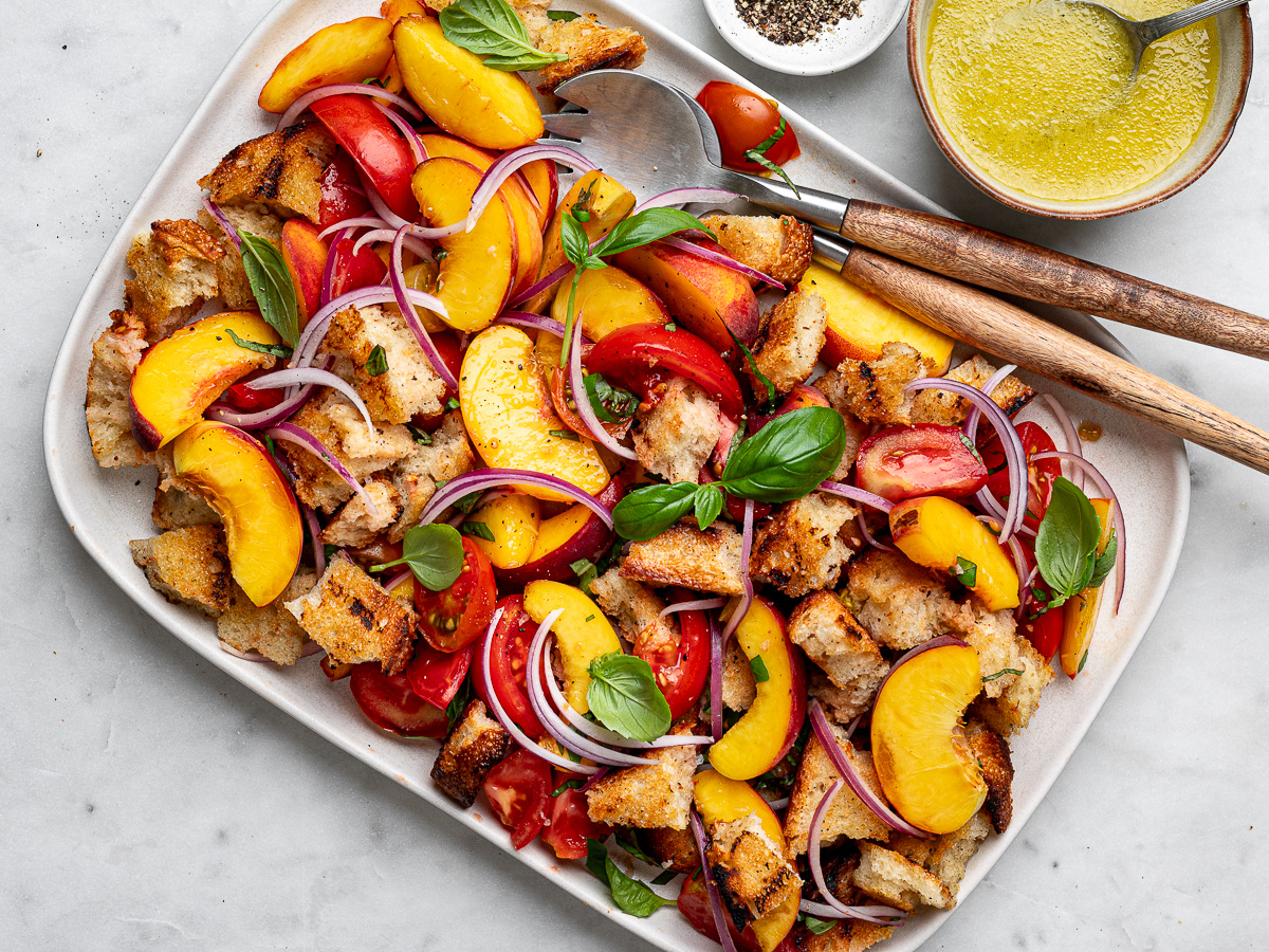 Peach panzanella salad layered on a large white platter with tomatoes, bread cubes, and red onion slices with salad tongs for serving.
