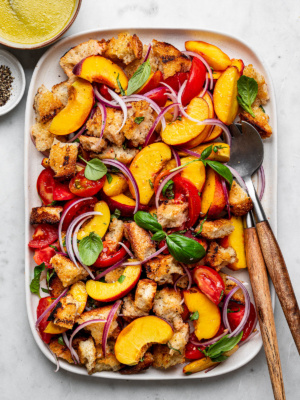 Peach panzanella salad layered on a large white platter with tomatoes, bread cubes, and red onion slices.