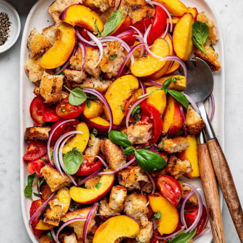 Peach panzanella salad layered on a large white platter with tomatoes, bread cubes, and red onion slices.