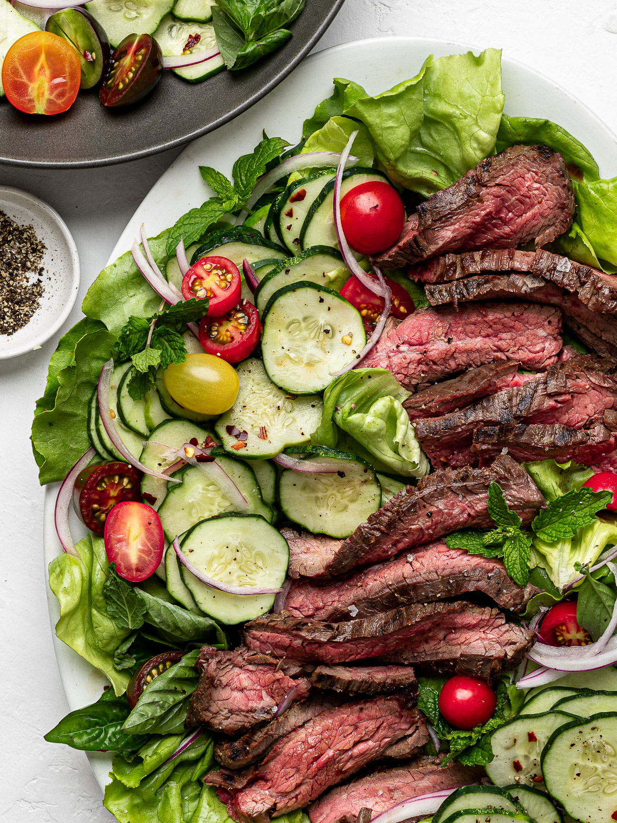 Close up of Slices of flank steak topped on a bed of lettuce with sliced cucumbers and cherry tomatoes.
