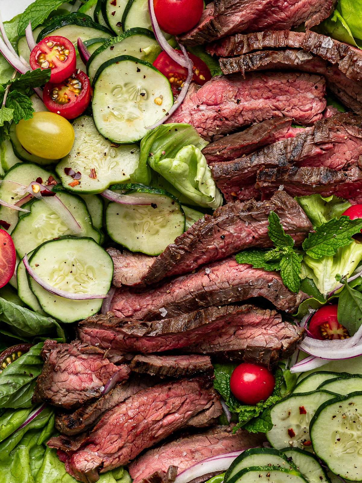 Close up of slices of flank steak topped on a bed of lettuce with sliced cucumbers and cherry tomatoes.