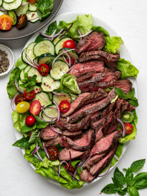 Slices of flank steak topped on a bed of lettuce with sliced cucumbers and cherry tomatoes.