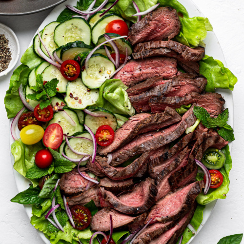 Slices of flank steak topped on a bed of lettuce with sliced cucumbers and cherry tomatoes.