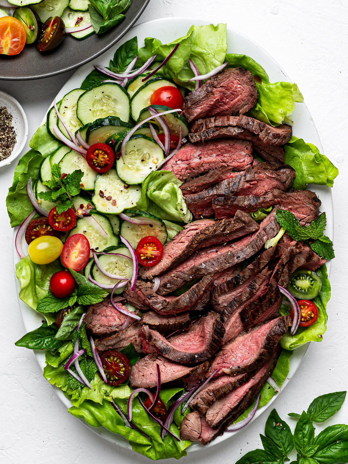 Slices of flank steak topped on a bed of lettuce with sliced cucumbers and cherry tomatoes.