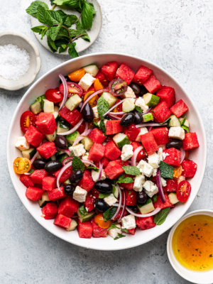 Watermelon salad served in white shallow bowl with fresh mint and dressing in bowls on the side.