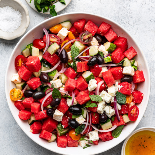Watermelon salad served in white shallow bowl with fresh mint and dressing in bowls on the side.