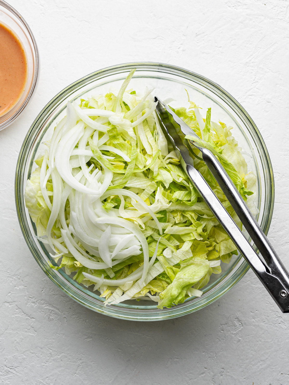 Lettuce-onion salad in a glass bowl with tongs and burger sauce bowl on the side.