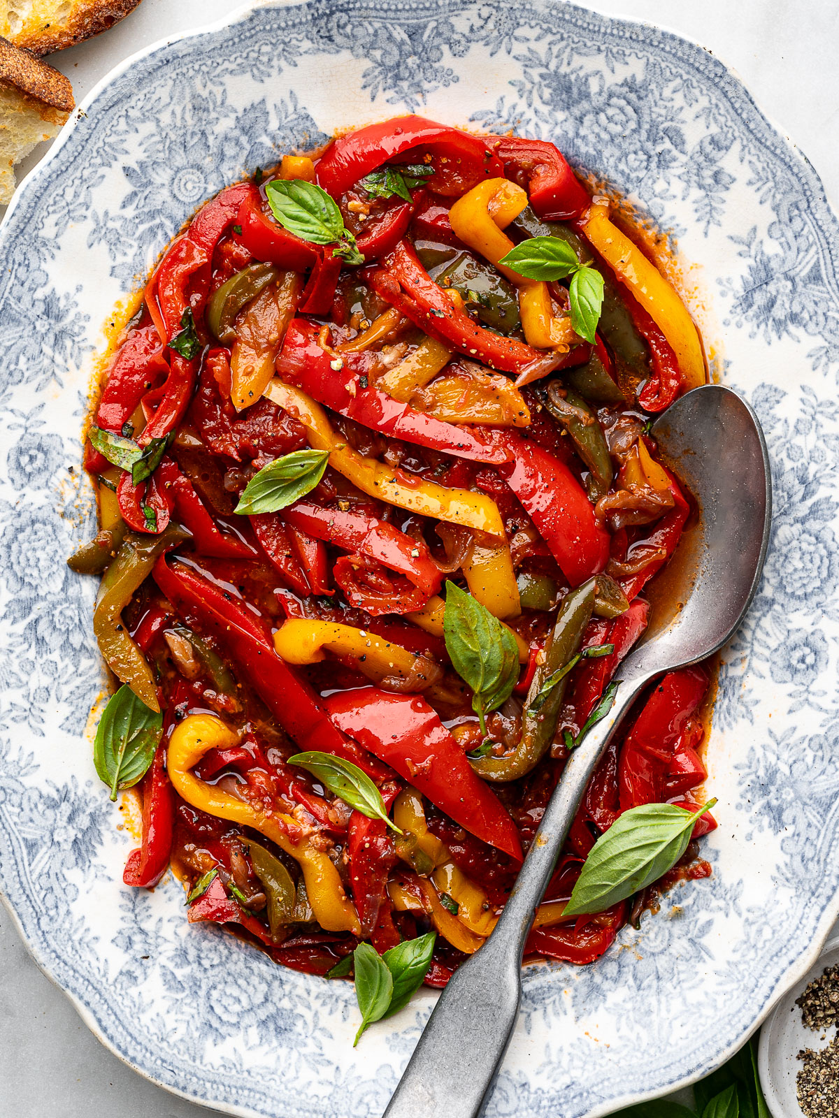 Close up of Peperonata (stewed peppers) served on a platter with a serving spoon and crusty bread on the side.