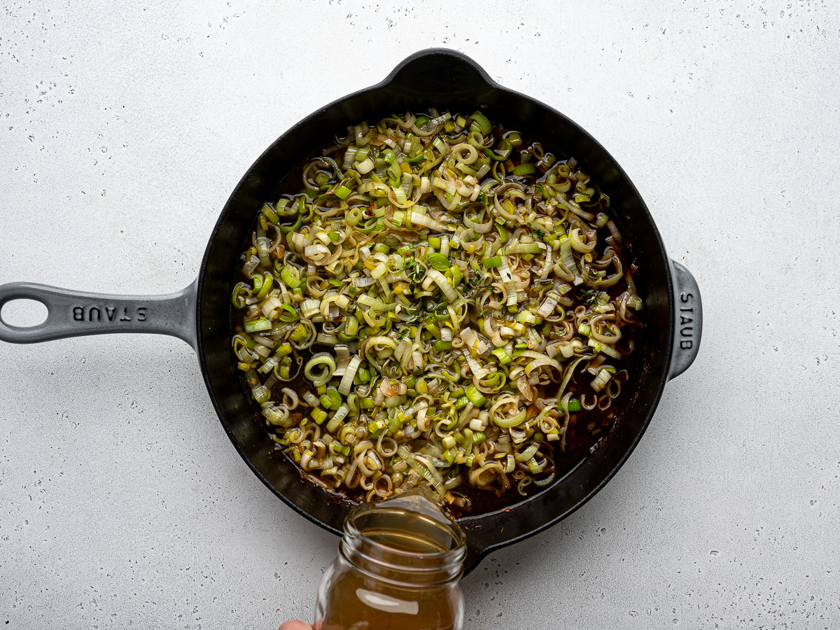 Adding chicken broth to sautéed leeks in skillet. 