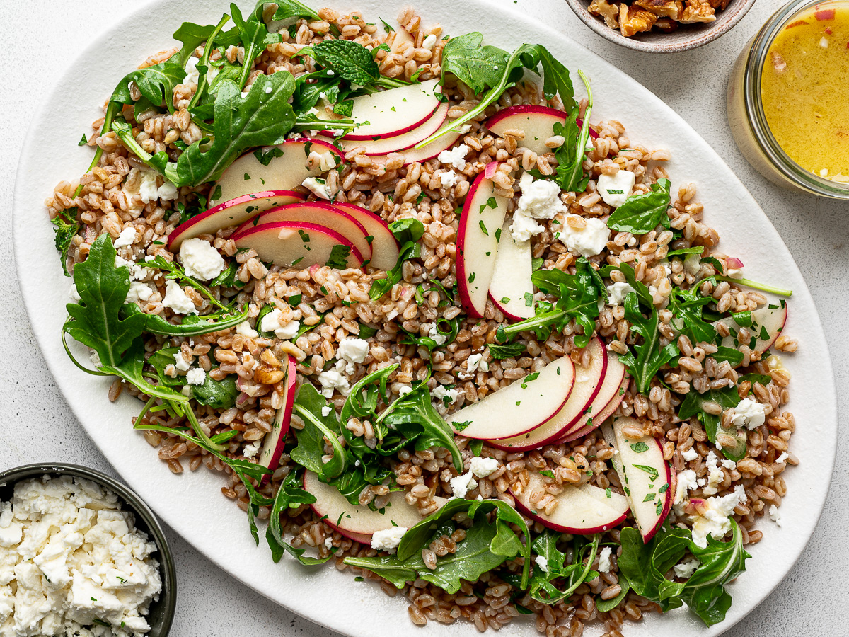 Farro Arugula Salad topped with apples, walnuts, feta, and herbs served on a white platter with jar of dressing, feta, and walnuts on the side.