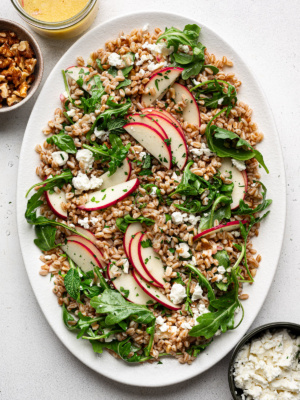 Farro Arugula Salad topped with apples, walnuts, feta, and herbs served on a white platter with jar of dressing, walnuts, and feta cheese on the side