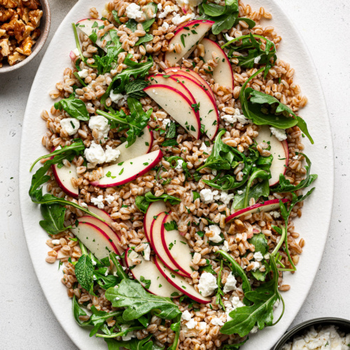 Farro Arugula Salad topped with apples, walnuts, feta, and herbs served on a white platter with jar of dressing, walnuts, and feta cheese on the side