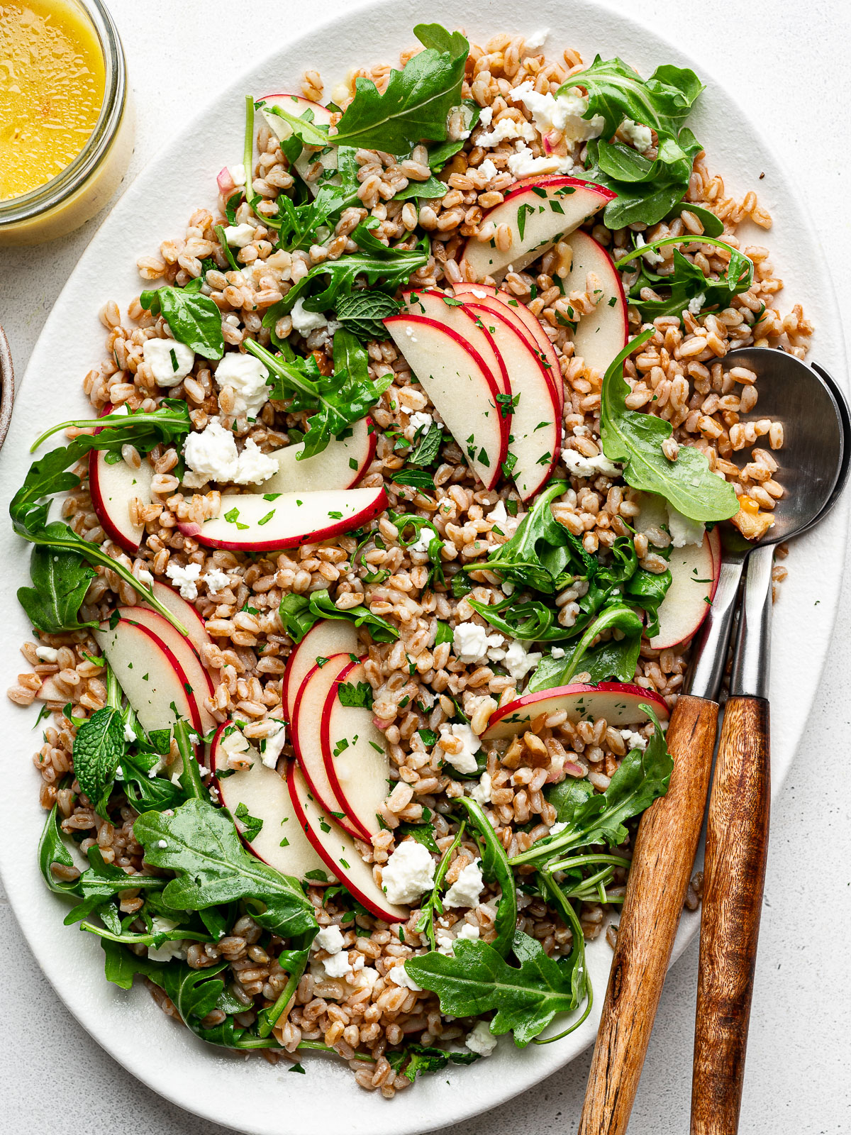 Farro Arugula Salad topped with apples, walnuts, feta, and herbs served on a white platter with jar of dressing on the side.