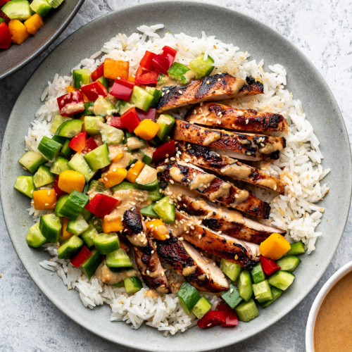 A grey plate with slices of Sesame Ginger Chicken on a bed of rice topped with mango-cucumber salad.