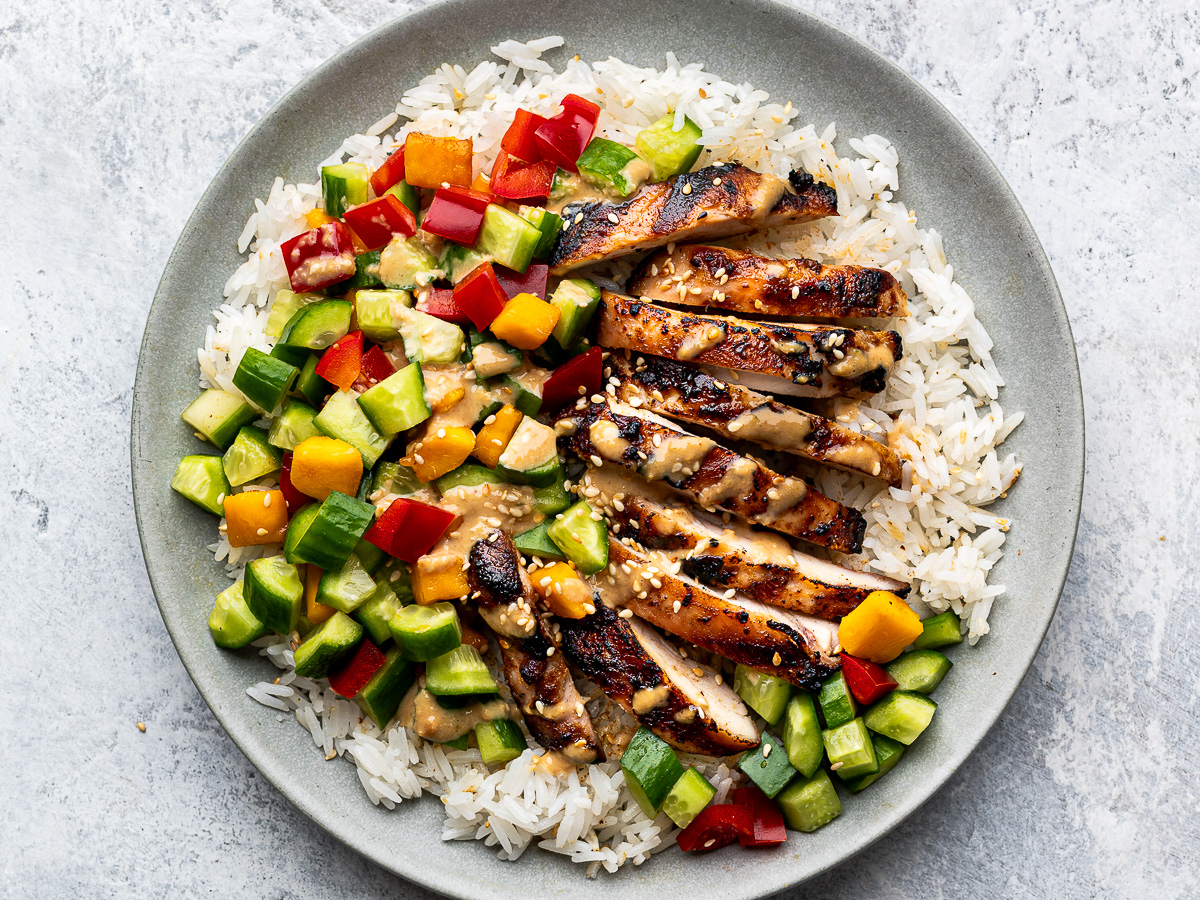 Close up of a grey plate with slices of Sesame Ginger Chicken on a bed of rice topped with mango-cucumber salad.