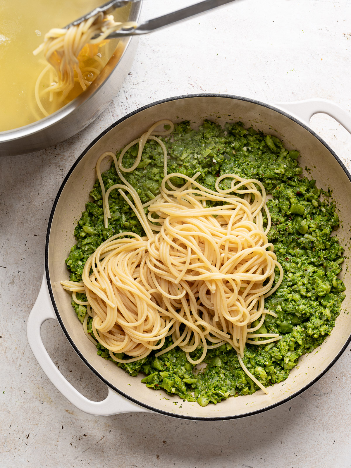 Scooping out cooked spaghetti from pot and adding to smashed broccoli in skillet. 