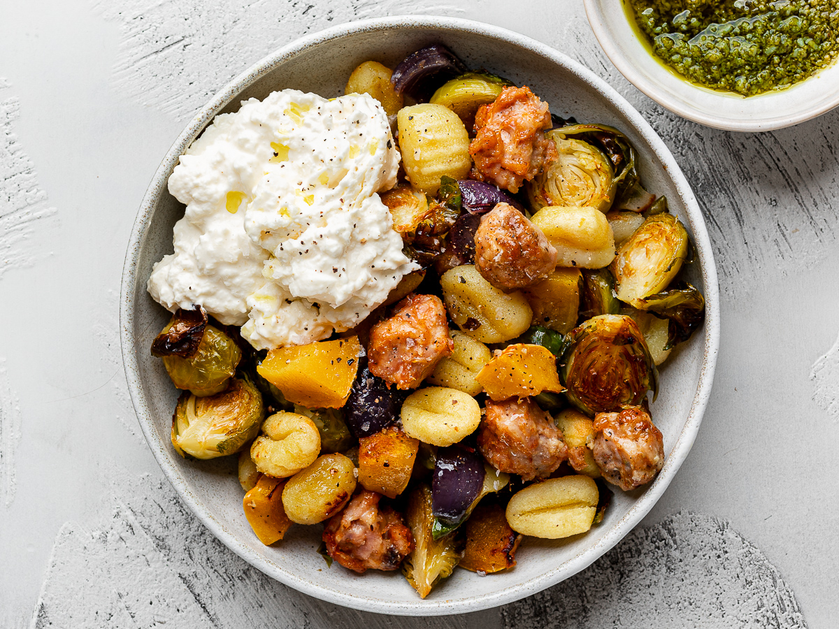 Roasted sheet pan gnocchi with Brussels sprouts, sausage squash and red onion served in a white bowl topped with burrata. 