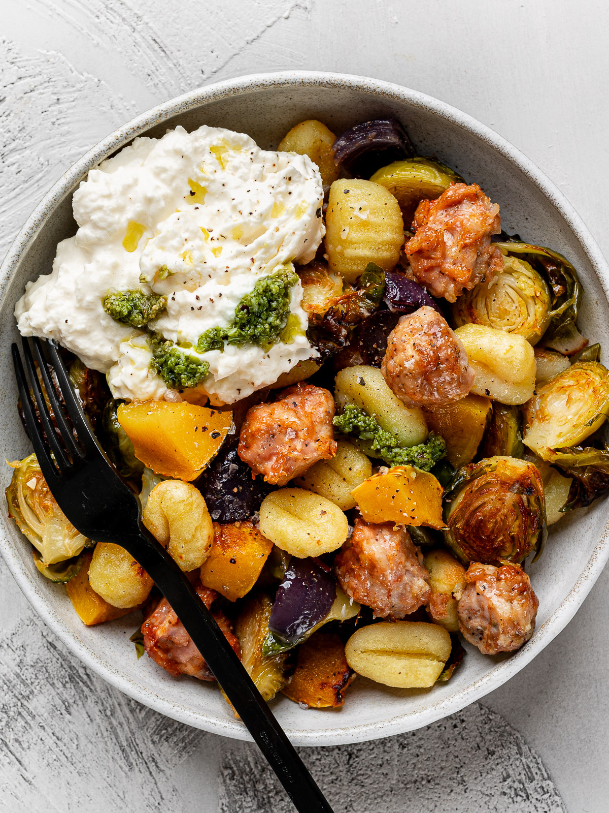 Roasted sheet pan gnocchi with Brussels sprouts, sausage squash and red onion served in a white bowl topped with burrata and a drizzle of pesto.  