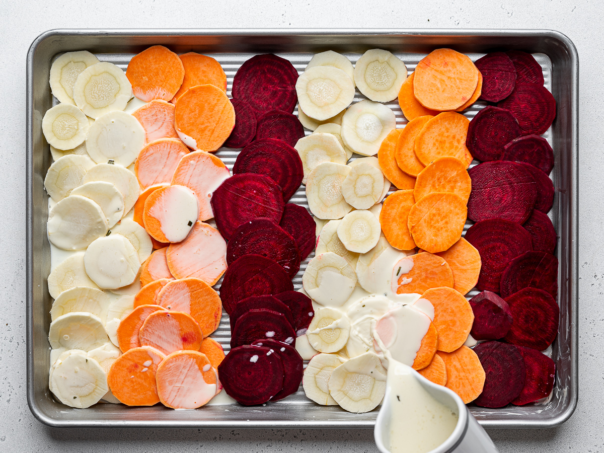 Pouring warmed cream mixture over sliced root vegetables in sheet pan.