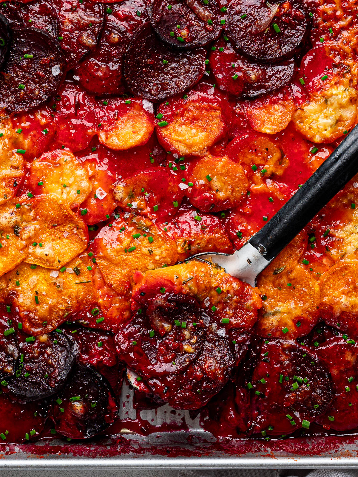 Close up of serving spoon scooping out a serving of root vegetable gratin from sheet pan. 