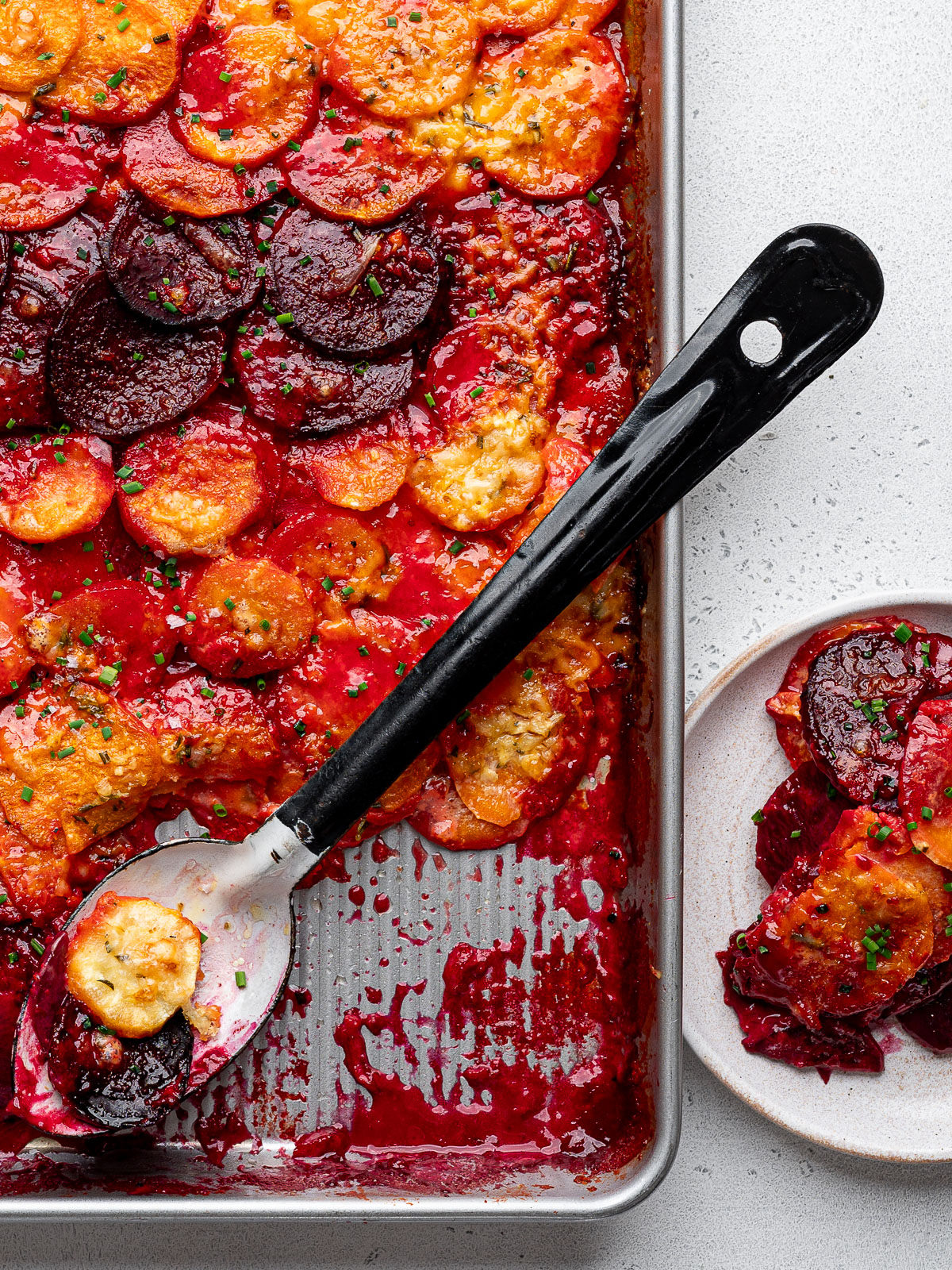 Serving spoon scooping out a serving of root vegetable gratin from sheet pan onto a small white plate.