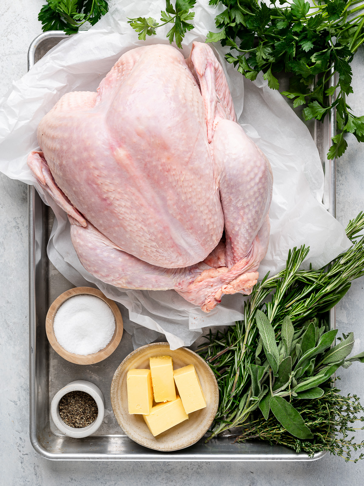 Recipe ingredients - raw turkey, herbs, butter, salt and pepper - prepared on a sheet pan.