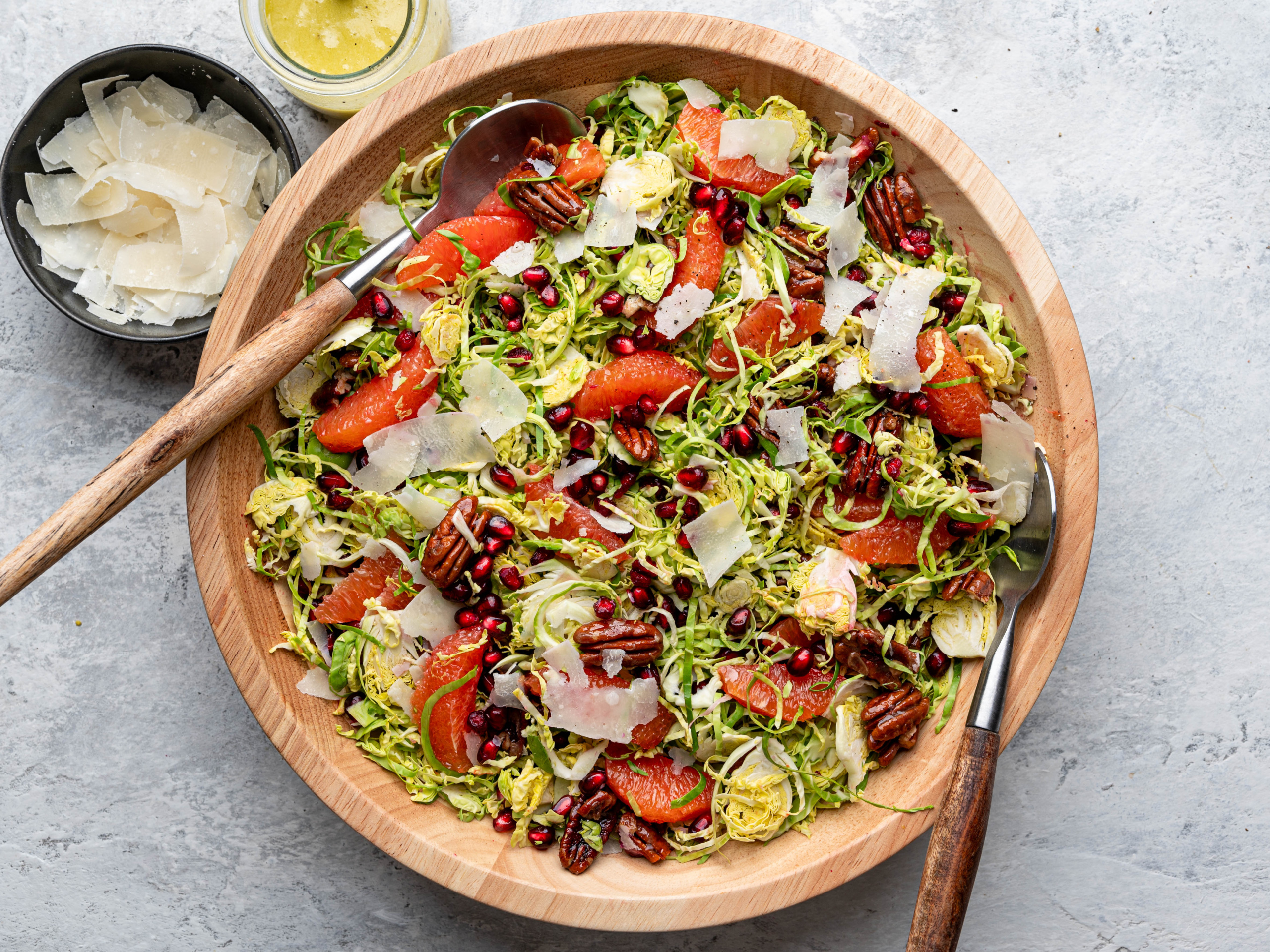 Tossed shaved Brussels sprouts salad in large wooden with vinaigrette and shaved parmesan cheese on the side.
