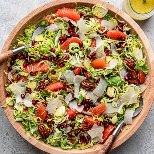 Tossed shaved Brussels sprouts salad in large wooden with vinaigrette in small jar on the side.